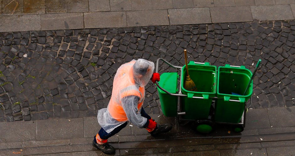 A person dressed in a gray hoodie, orange safety vest, and red gloves is operating a green cleaning cart with three compartments on a paved sidewalk. The cart contains cleaning tools such as a broom, 