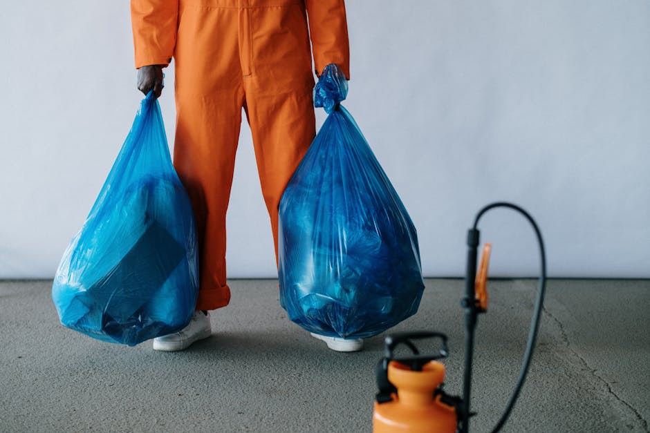 A person dressed in a gray hoodie, orange safety vest, and red gloves is operating a green cleaning cart with three compartments on a paved sidewalk. The cart contains cleaning tools such as a broom, 