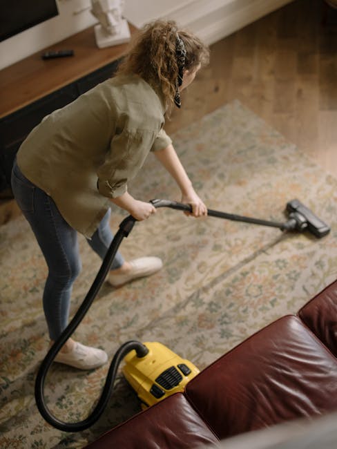 A woman with curly hair wearing a beige jacket, blue jeans, and white trainers is performing surface cleaning using a vacuum cleaner with a hose attachment on a large, patterned area rug in a living r
