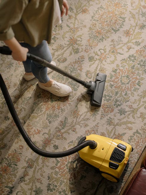 A woman with curly hair wearing a beige jacket, blue jeans, and white trainers is performing surface cleaning using a vacuum cleaner with a hose attachment on a large, patterned area rug in a living r